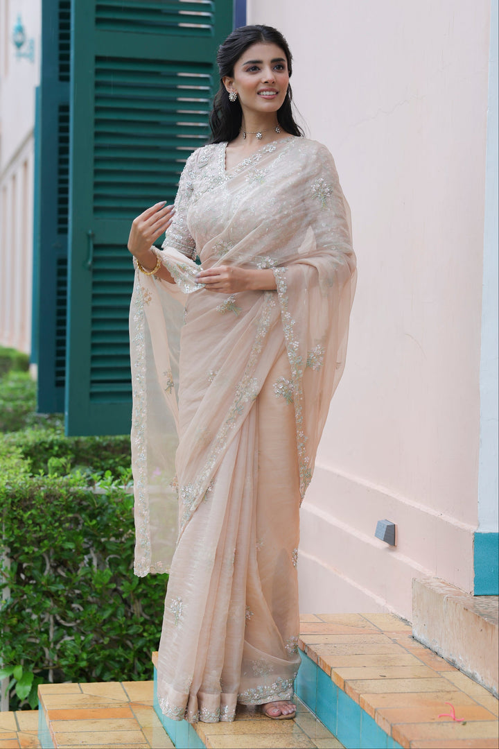 Woman in a beige saree standing outdoors near a building with green shutters.