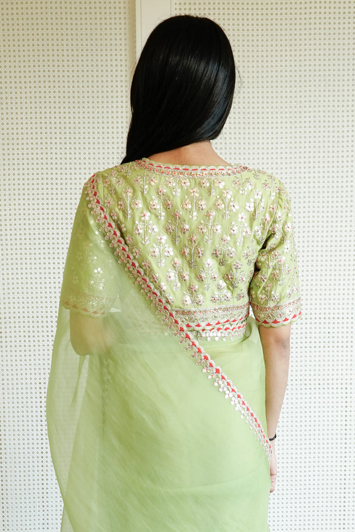 Person wearing a light green saree with intricate patterns, standing against a textured white wall.